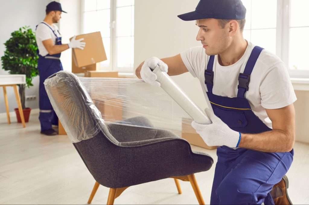 Canal Fulton mover with blue overalls and white gloves wraps a chair in plastic wrap while another mover moves boxes in the background. 
