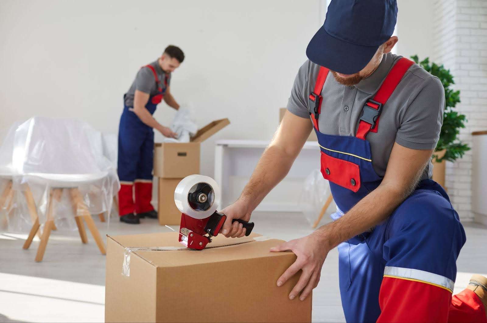 Mover in blue and red overalls taping up a box while another Kent Ohio mover packs a box in the background.